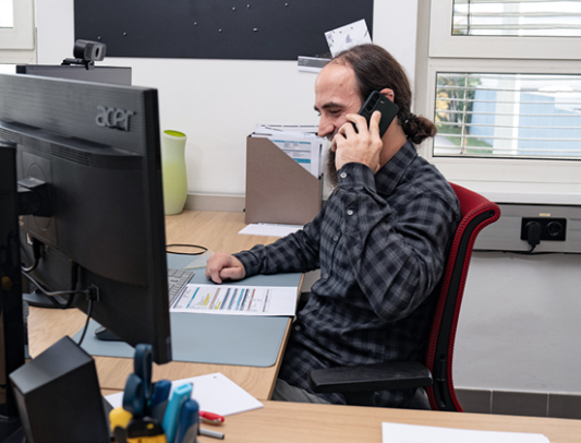 Mitarbeiter sitzt lächelnd am Schreibtisch im Büro hinter zwei Monitoren und telefoniert mit einem Handy